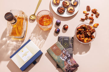 Flat lay of a Cocoa Dolce Monthly Chocolate Escape spread with handcrafted chocolate Moments, nuts, chocolate bars, and a glass of whiskey.