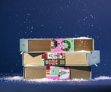 Stack of wrapped Cocoa Dolce holiday gift boxes in the snow, styled as a build-your-own chocolate advent calendar.
