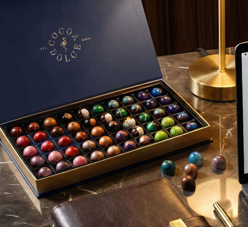 An open gold box of colorful hand-painted Cocoa Dolce chocolates on a table.