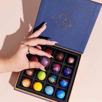 A hand lifting a hand-painted chocolate from an open navy Cocoa Dolce corporate gift box.
